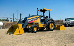 John Deere 210LEP Skip Loader parked on a dirt lot with construction vehicles in the background under a clear blue sky.