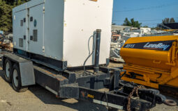 The image shows a towable Multiquip 180KVA generator mounted on a heavy-duty dual-axle trailer. The generator housing is white, with ventilation grilles and latches on the sides for access. The trailer has a black frame and it includes a hitch setup for towing, complete with a chain and heavy-duty hook. In the background, a yellow LeeBoy 8515 machine is visible, along with other industrial equipment. The scene appears to be in an outdoor yard or equipment lot.