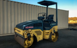 Bomag BW120AD-4 Vibratory Tandem Roller with Smooth Drum, featuring a compact design and an open canopy, parked on a dirt lot in front of a beige shipping container at sunset.